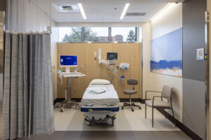 An empty patient recovery room is lit by clerestory windows. A white bed is at the center surrounded by various monitoring equipment.