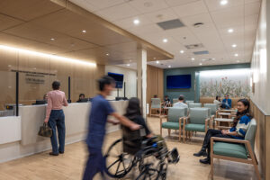 A health clinic waiting room. In the foreground, a person in blue scrubs pushes another person in a wheelchair.