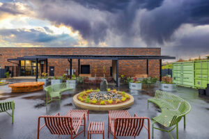 A low brick building has a dark metal canopy structure in front of it. Several colorful chairs and benches are arranged around a circular flower bed in front of the canopy.