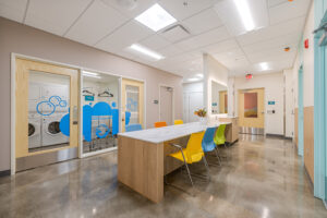Colorful chairs are lined up on either side of a counter island in a brightly lit room. Laundry facilities are visible beyond glass doors and windows decorated with blue bubble graphics.