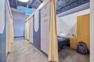 A curtain is drawn back to reveal a small partitioned room within a congregate shelter.
