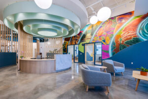 A lobby with a central wooden reception desk has a large circular fixture overhead. A colorful mural decorates the far wall.