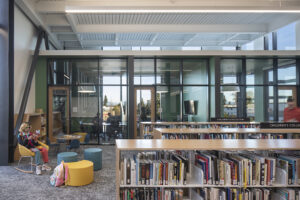 Rows of bookshelves labeled "Children's Collection". A small seating area is off to the left where a student sits in a rocking chair and reads a book.