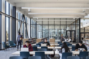 College students study at computer desks in a large, high-ceilinged space with tall floor-to-ceiling windows.