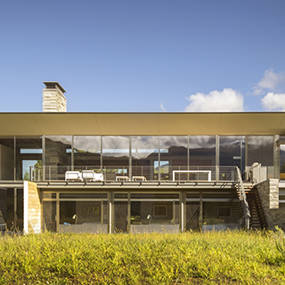 A two-story house with a wide overhanging roof sits in a meadow with a blue sky overhead.