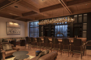 A warmly lit bar with a row of empty chairs in front of it. There is a large light fixture hanging over the bar that is made of many interlocked deer antlers.