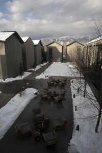 A snowy courtyard with several fire pits is surrounded by multiple tall, pointy-roofed gray buildings.