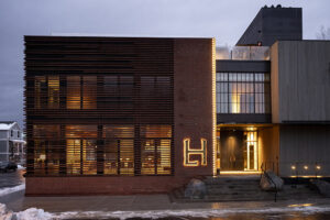 A brick and concrete building with a warmly glowing entry features a large, illuminated "LH" sign beside the front door.