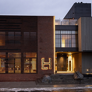 A brick and concrete building with a warmly glowing entry features a large, illuminated "LH" sign beside the front door.