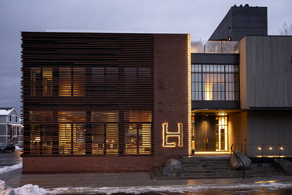Larch-House-Exterior A brick and concrete building with a warmly glowing entry features a large, illuminated "LH" sign beside the front door.