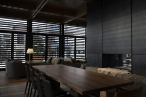 A long, dark wood table in front of a black fireplace is in the center of the frame. In the background, chairs for a restaurant line along the window which is darkened with horizontal slats.