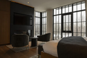 A hotel room with two chairs in front of a black fireplace and a bed in the foreground. Tall windows line the back wall of the room, showing a snowy landscape outside.