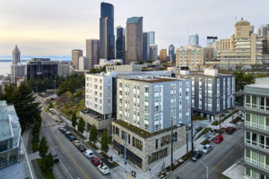 A large rectangular apartment building sits at the corner of a street intersection. Skyscrapers rise in the background.