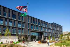 A large, rectangular brick building with many windows. A flagpole and entry plaza are in the foreground.