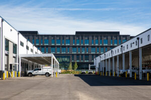 A large brown building with many windows in the background is framed on either side by lower, white shop buildings full of vehicles in the foreground.