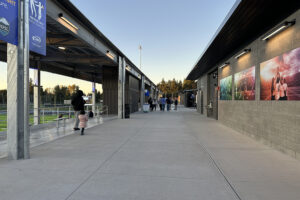 A wide, uncovered walkway between the back side of a stadium structure and a brick restroom/concessions building.
