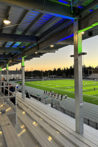The underside of a metal stadium roof with green and blue lights, overlooking bleachers, a running track and athletic field.