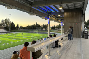 The underside of a metal stadium roof with green and blue lights, overlooking bleachers, a running track and athletic field.