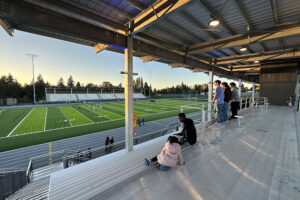 The underside of a metal stadium roof with green and blue lights, overlooking bleachers, a running track and athletic field.