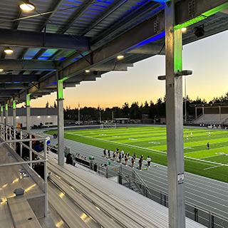 The underside of a metal stadium roof with green and blue lights, overlooking bleachers, a running track and athletic field.
