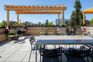 People relax on a rooftop patio with planters around the edge and a wooden pergola overhead.