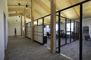A well-lit wood ceiling covers a cubicle room divider wall. A person walks down the hallway in the background.