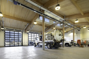 A large utility vehicle is inside a wood-framed garage. A tall wood ceiling is lit by clerestory windows and windowed garage doors.