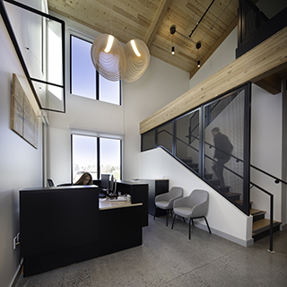 A woman sits at a reception desk in a tall, brightly lit room. A person walks up a partially screened staircase in the background.